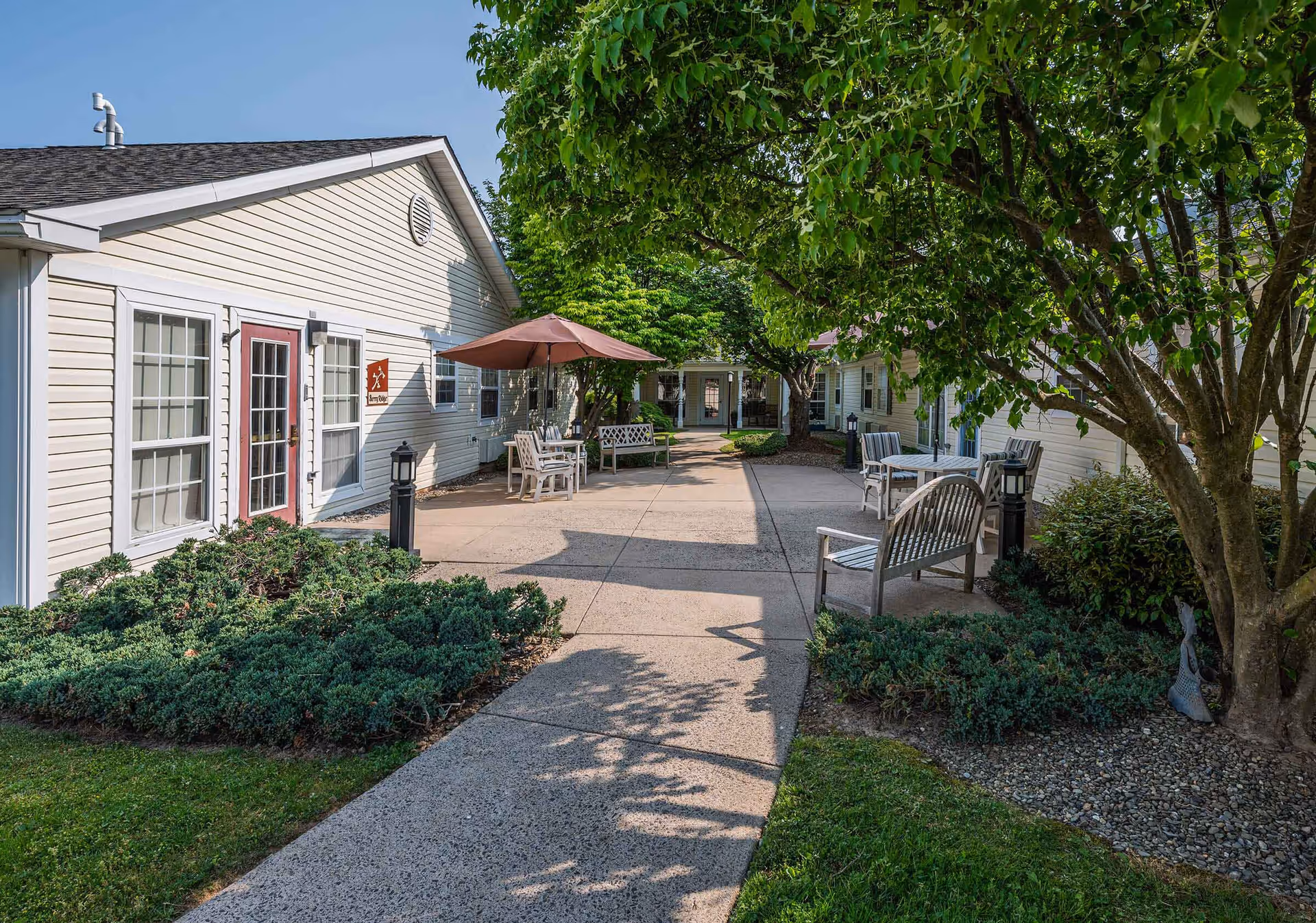Outdoor courtyard area at Arden Courts - ProMedica Memory Care Community (Yardley) featuring a paved walkway, benches, tables with umbrellas, surrounded by green bushes and trees, and light-colored buildings with windows and doors.