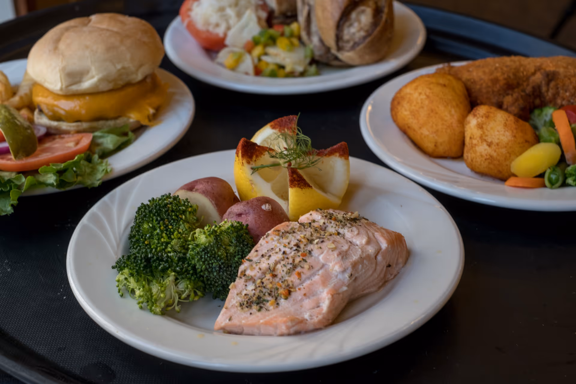 A close-up of four plates of food on a black tray. The front plate has a serving of seasoned salmon, broccoli, red potatoes, and a lemon garnish. The other plates include a cheeseburger with lettuce, tomato, and pickle, a sandwich with a side of mixed vegetables and potato salad, and a plate with fried food and mixed vegetables.
