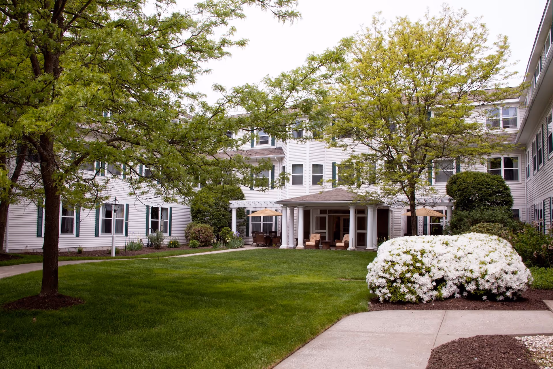 Outdoor courtyard area of a senior living facility with green grass, trees, white flowering bushes, and a three-story white building with multiple windows surrounding the courtyard. There are chairs and tables under a covered patio area.