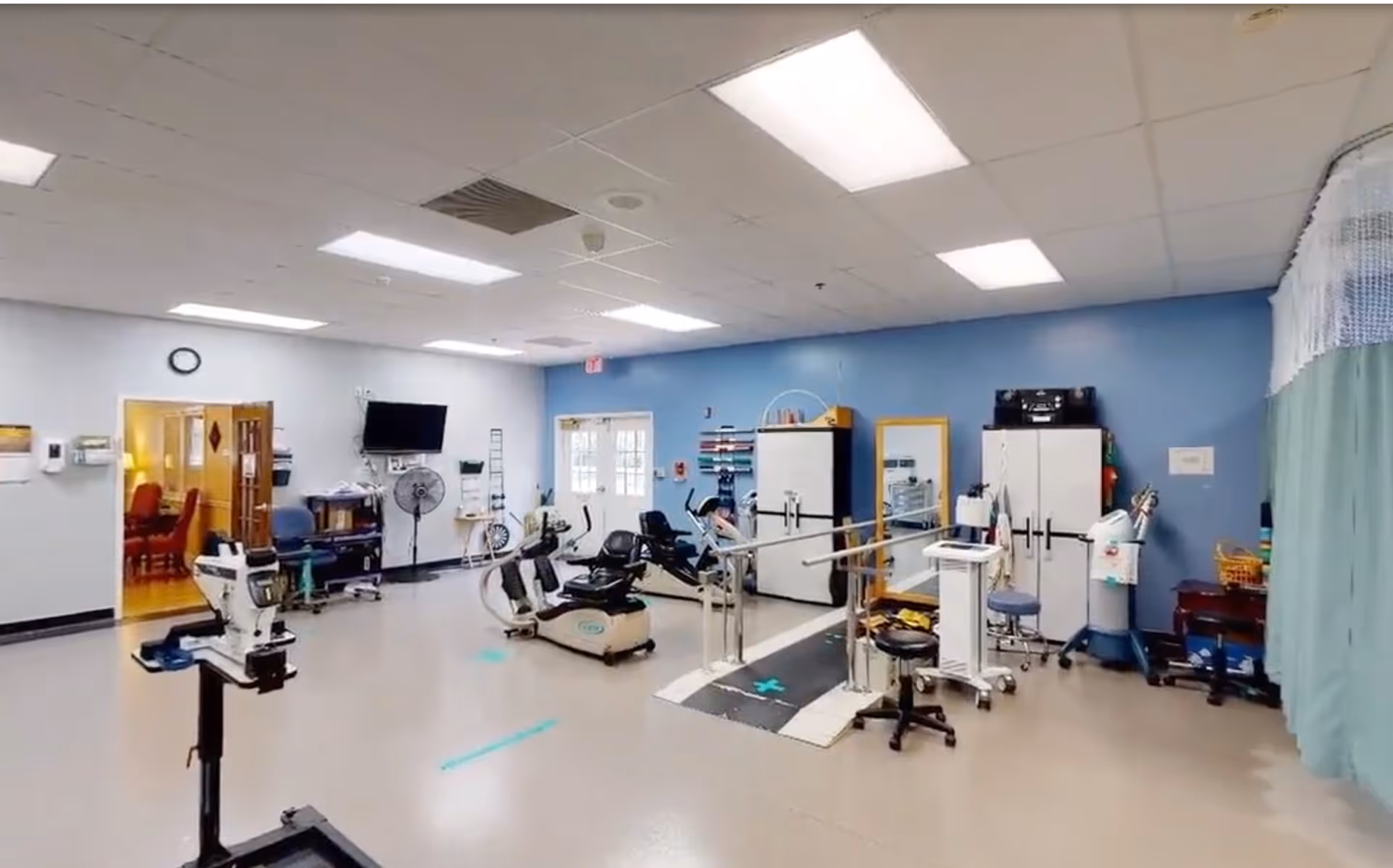 A spacious physical therapy or rehabilitation room with exercise equipment including stationary bikes and parallel bars for walking practice. The room has a blue accent wall with cabinets, a mirror, and various medical and therapy supplies. There is a doorway leading to another room with a dining area visible in the background.