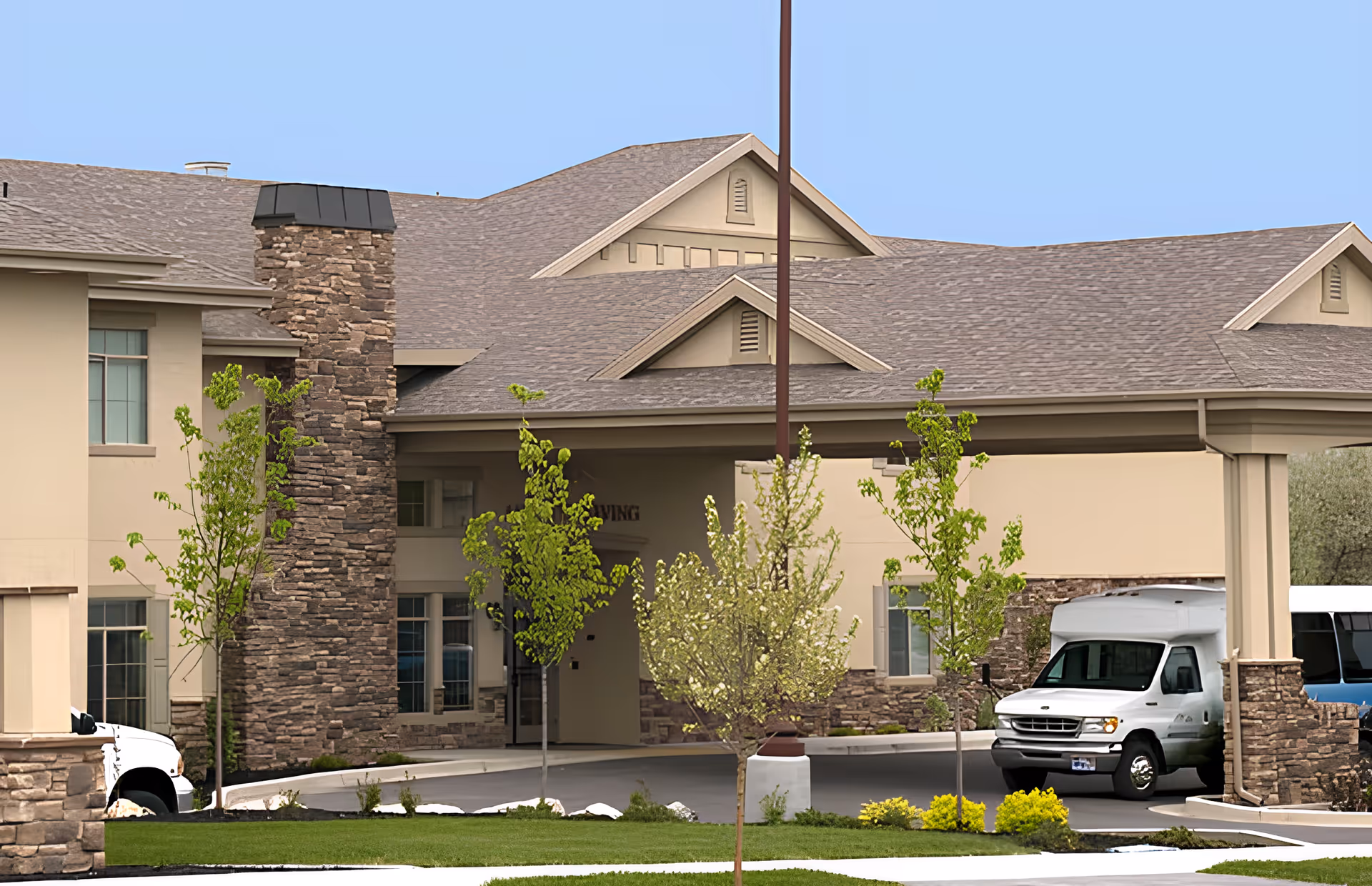 Exterior view of Carrington Court Assisted Living and Memory Care building entrance with a covered driveway, stone and beige walls, several small trees, and a white shuttle van parked under the entrance canopy.