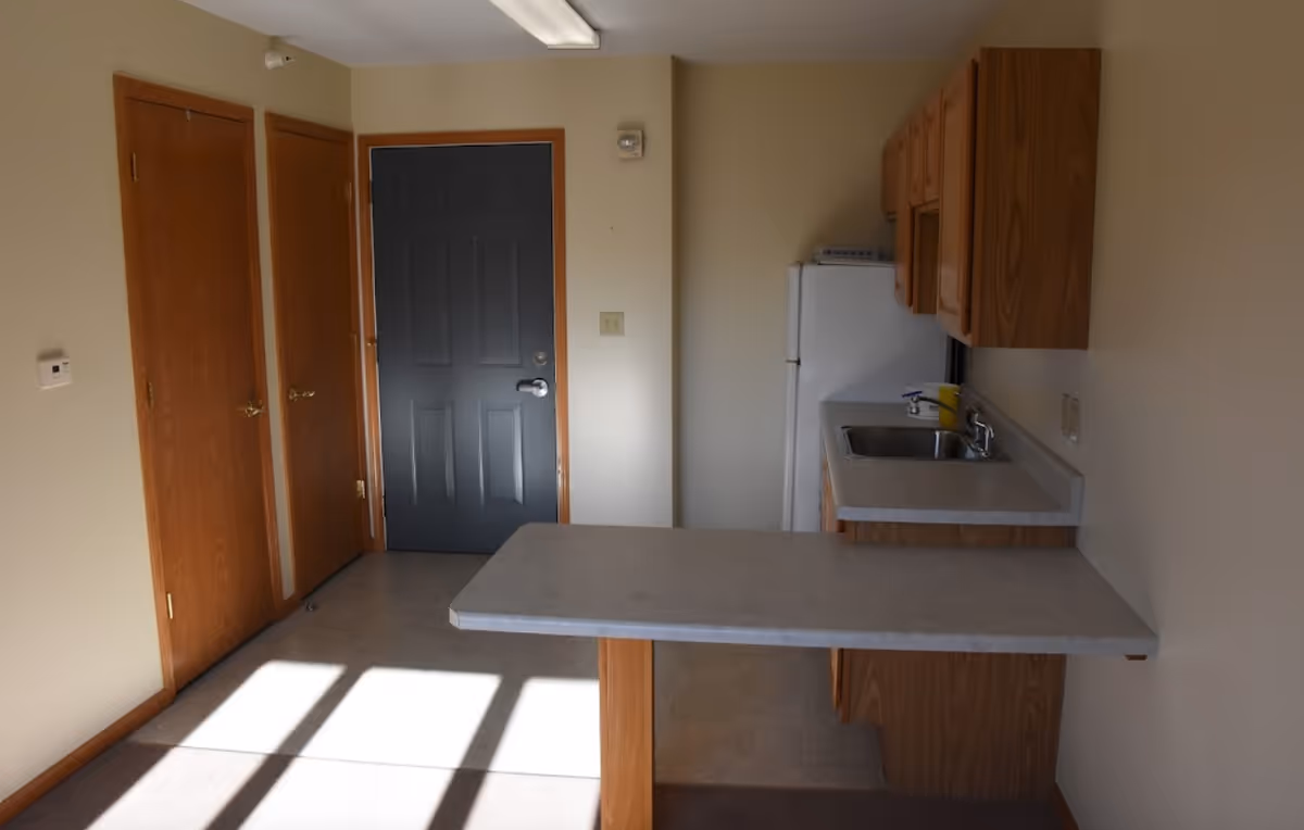 Small kitchenette with a laminate countertop peninsula, sink, upper wood cabinets, refrigerator and a closed gray entry door.