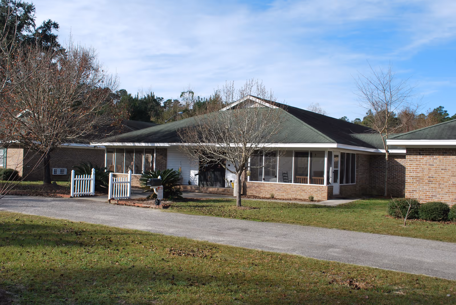Exterior view of a single-story brick and siding building with a green roof, surrounded by a lawn with leafless trees and shrubs under a partly cloudy sky.