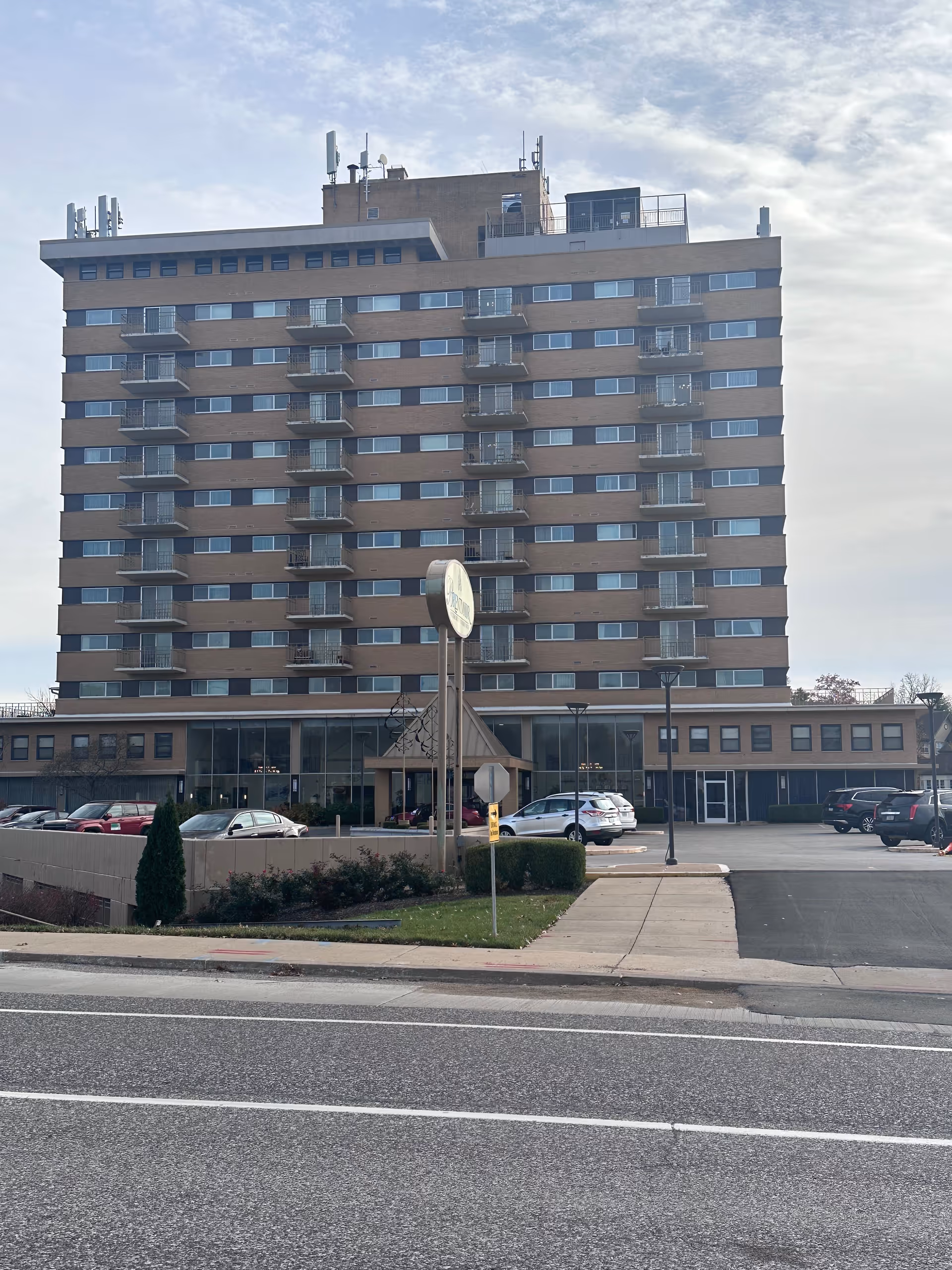 A multi-story building with numerous balconies and windows, identified as Brentmoor Retirement Community, viewed from across the street with a parking lot and cars in front. The sky is partly cloudy.