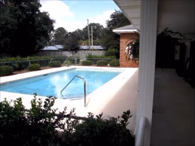 Outdoor swimming pool surrounded by a concrete deck with bushes and trees in the background, adjacent to a covered patio area with white pillars and a brick wall.