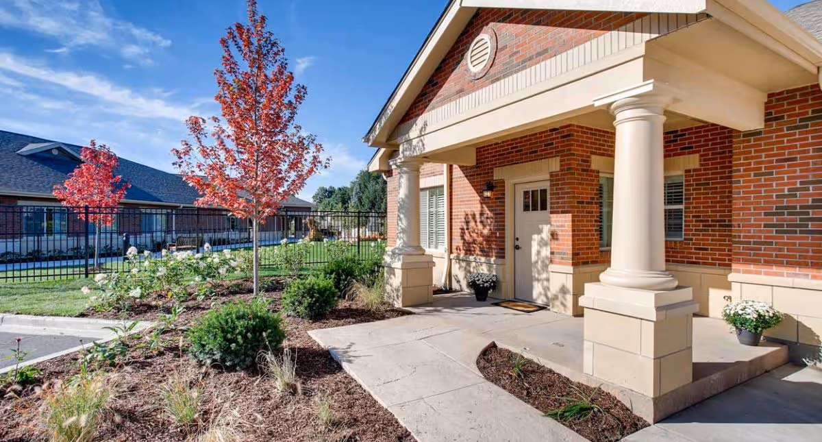 Exterior view of a brick building with white columns at the entrance, a concrete walkway, landscaped garden beds with shrubs and a tree with red leaves, and a fenced grassy area under a blue sky with some clouds.