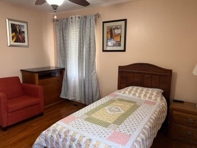 Single-occupancy bedroom with a wooden twin bed covered by a patchwork quilt, a red armchair, dresser and nightstand beside a curtained window.
