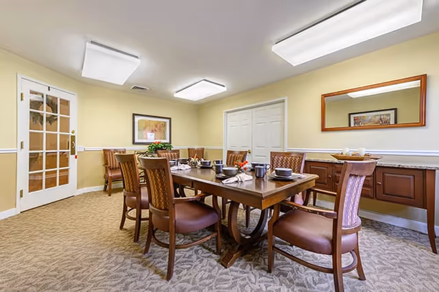 A dining room with a rectangular wooden table set for six people with plates, cups, and napkins. The room has beige walls, carpeted floor, a large mirror on one wall, a framed picture on another, and a wooden sideboard beneath the mirror. There is a door with glass panels on one side and two ceiling lights providing illumination.