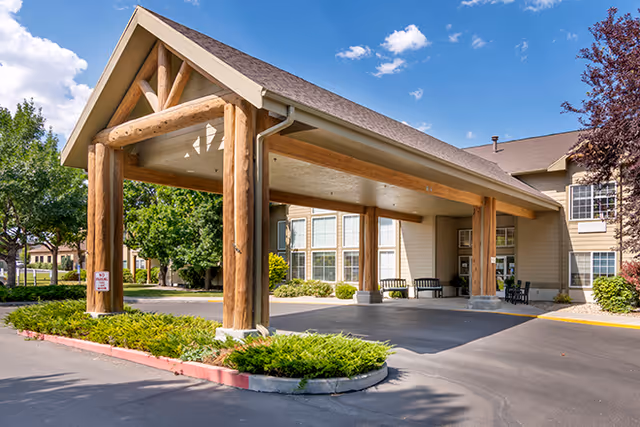 Entrance area of a senior living facility with a large covered driveway supported by wooden beams. The building has beige siding and multiple windows. There are benches near the entrance and landscaped greenery around the driveway under a partly cloudy blue sky.
