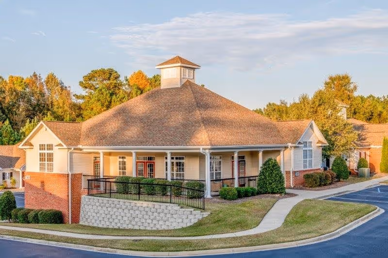 Exterior view of a single-story senior living facility building with beige siding and a brown shingled roof, surrounded by landscaped greenery and a paved driveway under a partly cloudy sky.