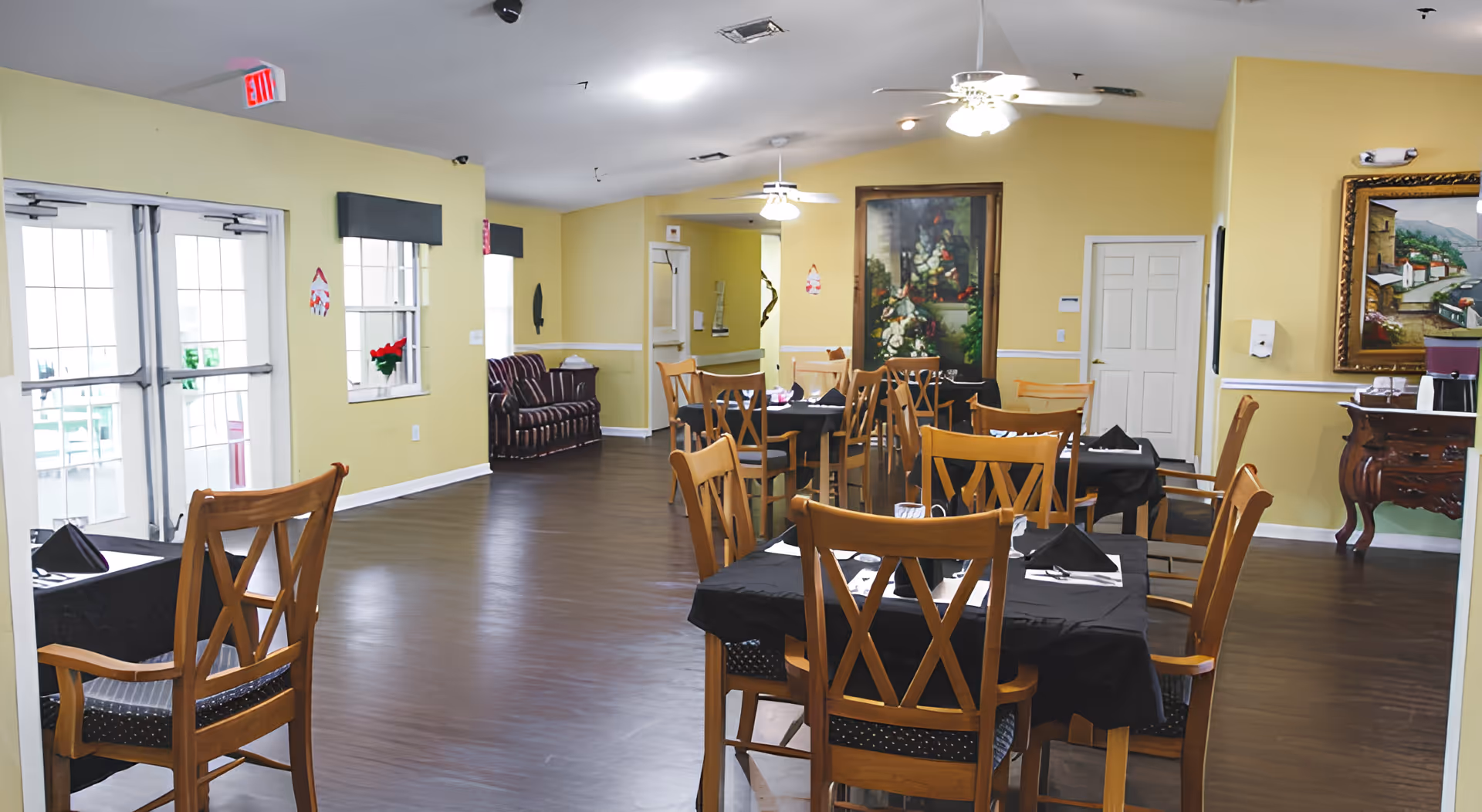 A dining room in a senior living facility with several wooden tables and chairs arranged neatly. The tables are covered with black tablecloths and set with napkins and utensils. The room has yellow walls, dark wood flooring, ceiling fans with lights, and decorative paintings on the walls. There is a striped couch along one wall and double glass doors leading outside.