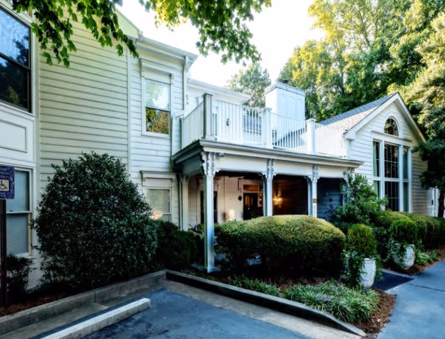 Front exterior of a two-story light-colored building with a covered porch, second-floor balcony, landscaped shrubs, and a small driveway.