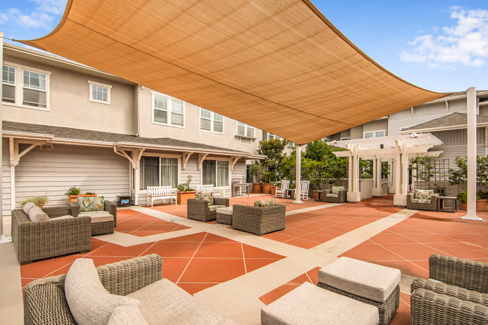 Outdoor courtyard with a large fabric shade sail, wicker lounge furniture, pergola, and a tiled patio beside a two-story building.