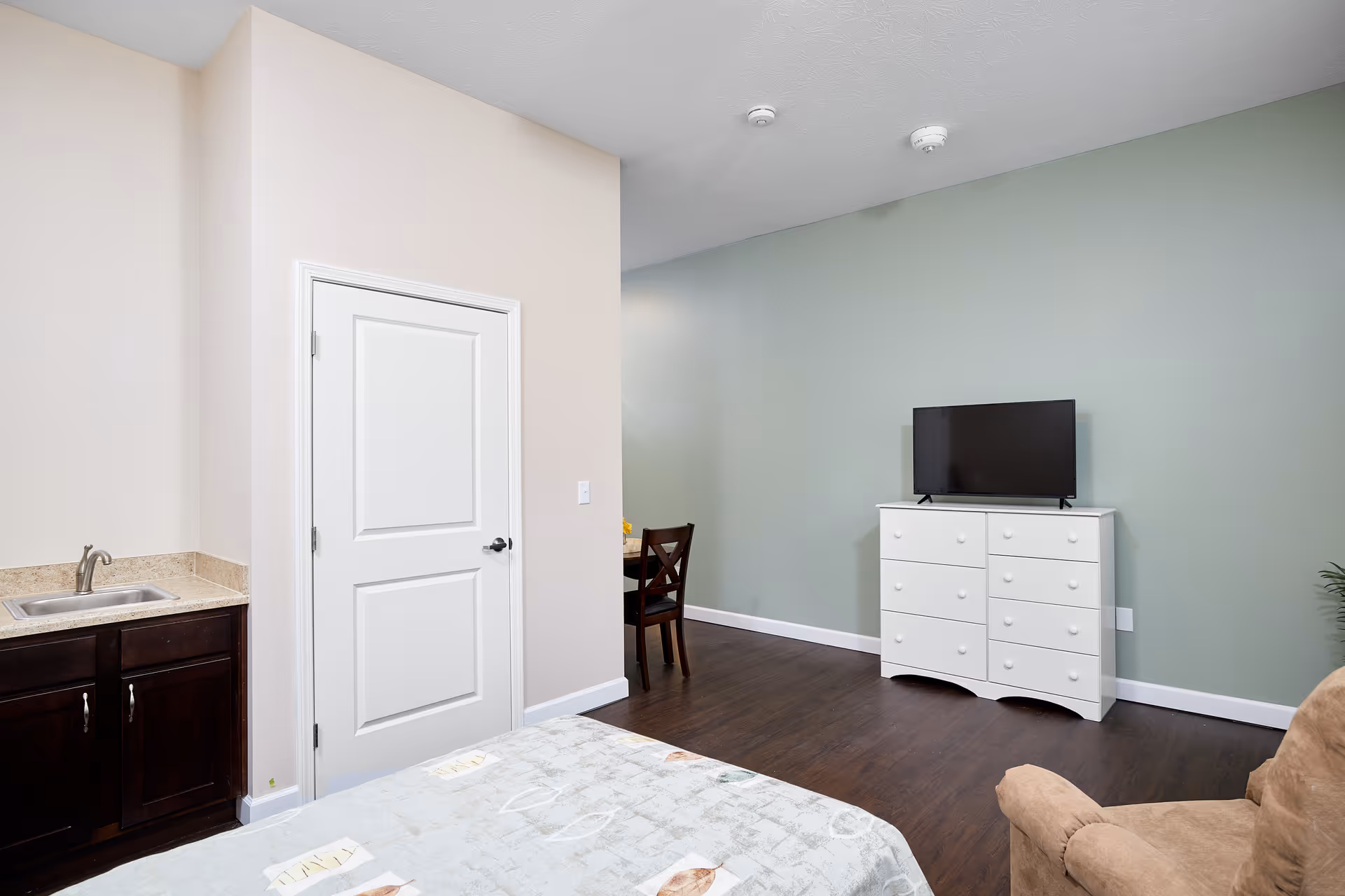 A room with a bed partially visible in the foreground, a small sink with dark cabinetry on the left, a white door in the center, a white dresser with a flat-screen TV on top against a green wall, a wooden chair and table in the background, and a beige armchair on the right.