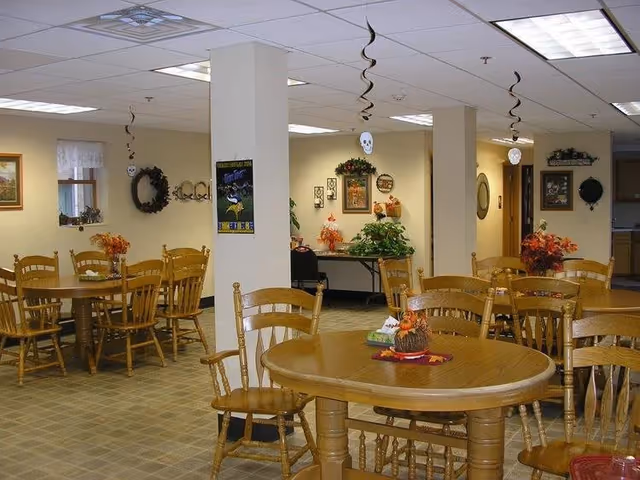 A dining area with multiple wooden tables and chairs arranged neatly. The room is decorated with autumn-themed centerpieces and hanging Halloween decorations such as skulls. The walls have framed pictures, wreaths, and plants, creating a cozy atmosphere.