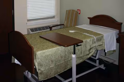 Senior living bedroom with adjustable bed, over-bed table, window blinds, and a chair.