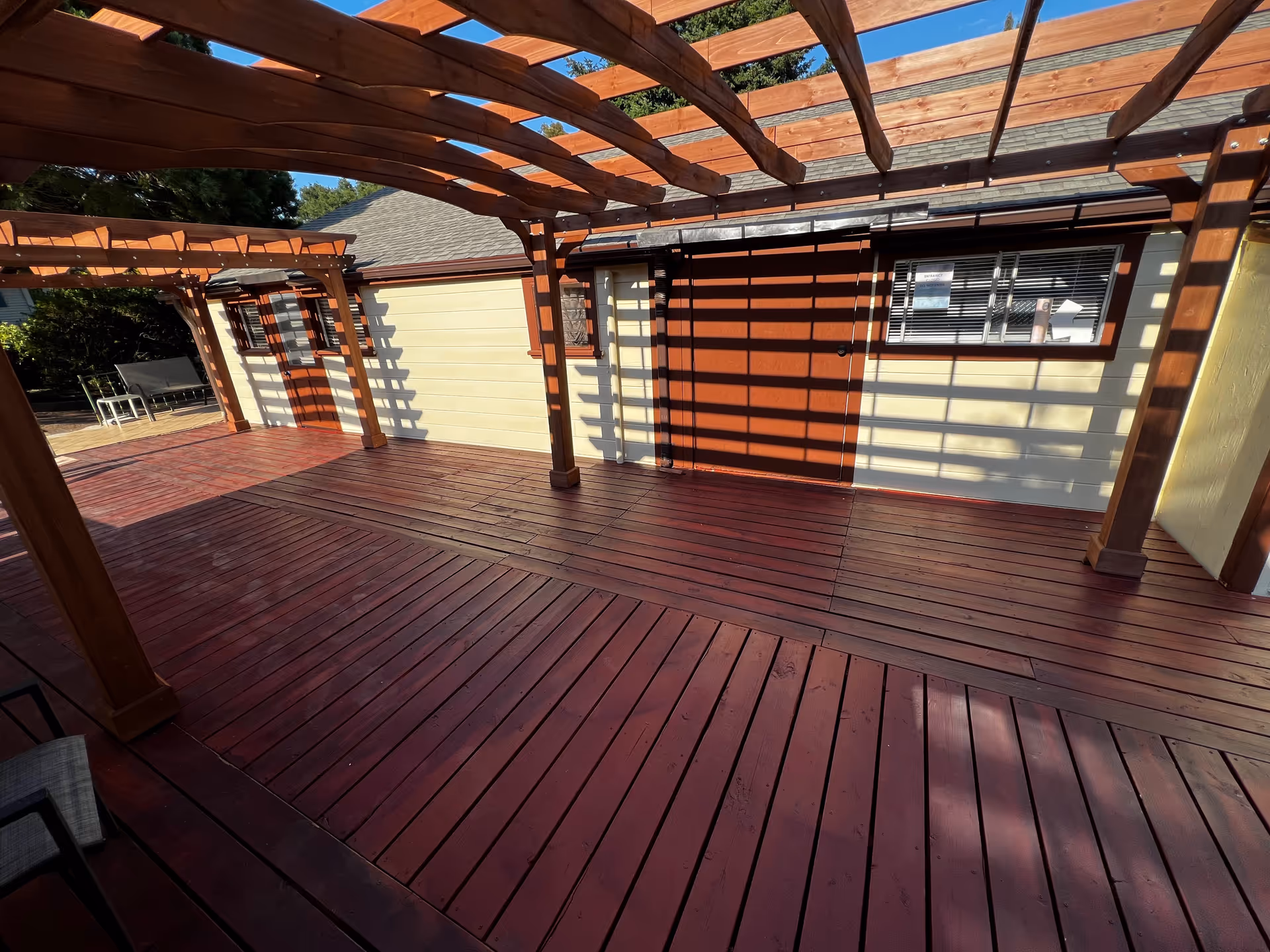 Outdoor wooden deck area with a pergola casting shadows on the floor and walls. The deck is adjacent to a building with cream-colored siding and a red door and window frame. There is outdoor seating visible in the background under another pergola.