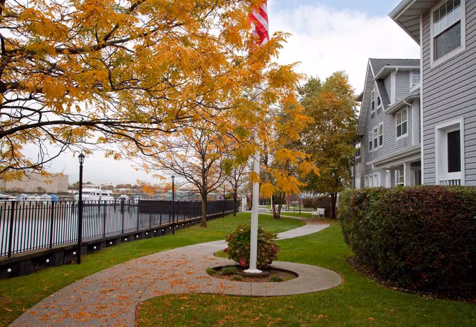 A winding concrete pathway lined with green grass and autumn trees with yellow leaves, running alongside a gray multi-story building and a black metal fence by the water. An American flag on a white flagpole is centered in the image.