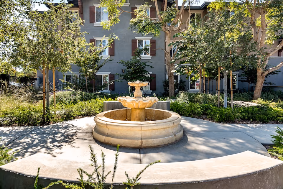 A stone tiered fountain in a landscaped courtyard with trees and a multi-story building facade in the background.
