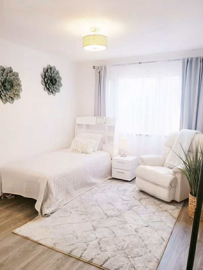Bright, minimalist bedroom with a twin bed, white nightstand and lamp, a plush recliner, decorative wall art and a large curtained window.