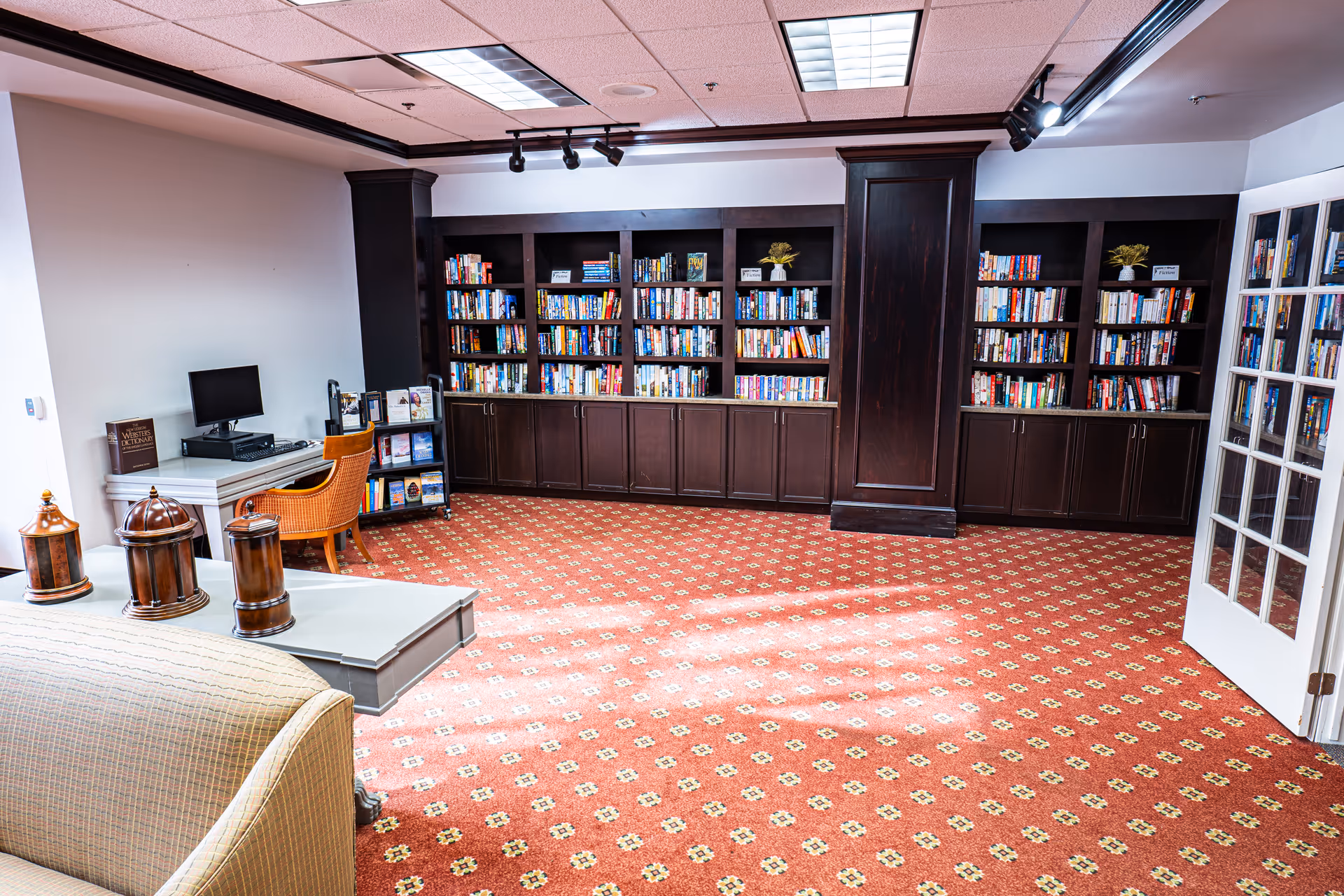 A cozy library room with dark wooden bookshelves filled with books along the back wall. There is a computer desk with a monitor and a wicker chair on the left side. The floor is covered with a red patterned carpet, and a beige sofa is partially visible in the foreground. The room has white walls and a ceiling with fluorescent lights and a track light fixture.
