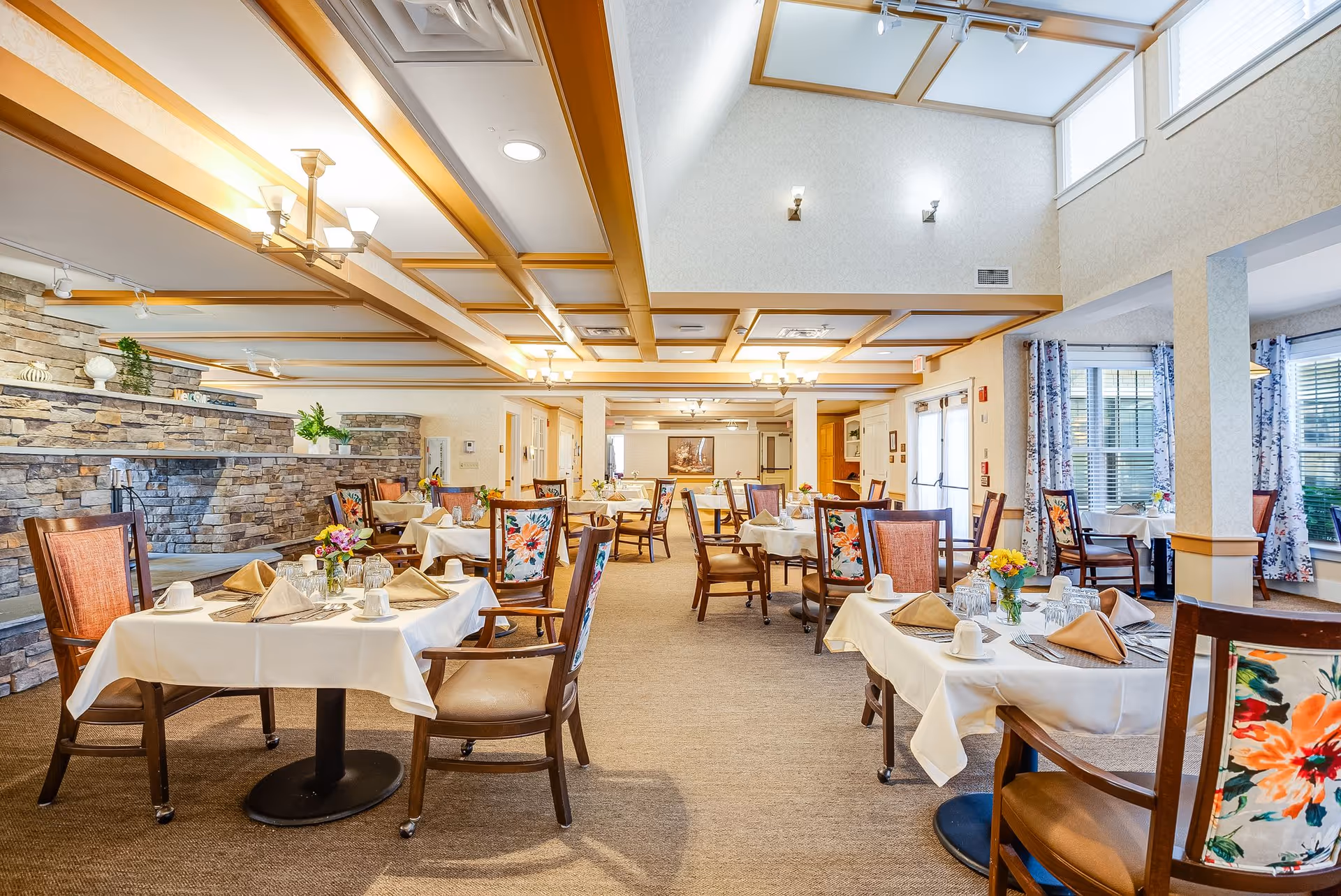 Spacious, well-lit dining room with neatly set tables, floral-upholstered chairs, and a stone accent wall.