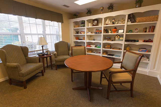 A cozy sitting area with two striped armchairs and one wooden chair around a round wooden table. Behind the table is a large built-in white bookshelf filled with various decorative items and books. A window with blinds and a small table with a lamp and a phone are on the left side.