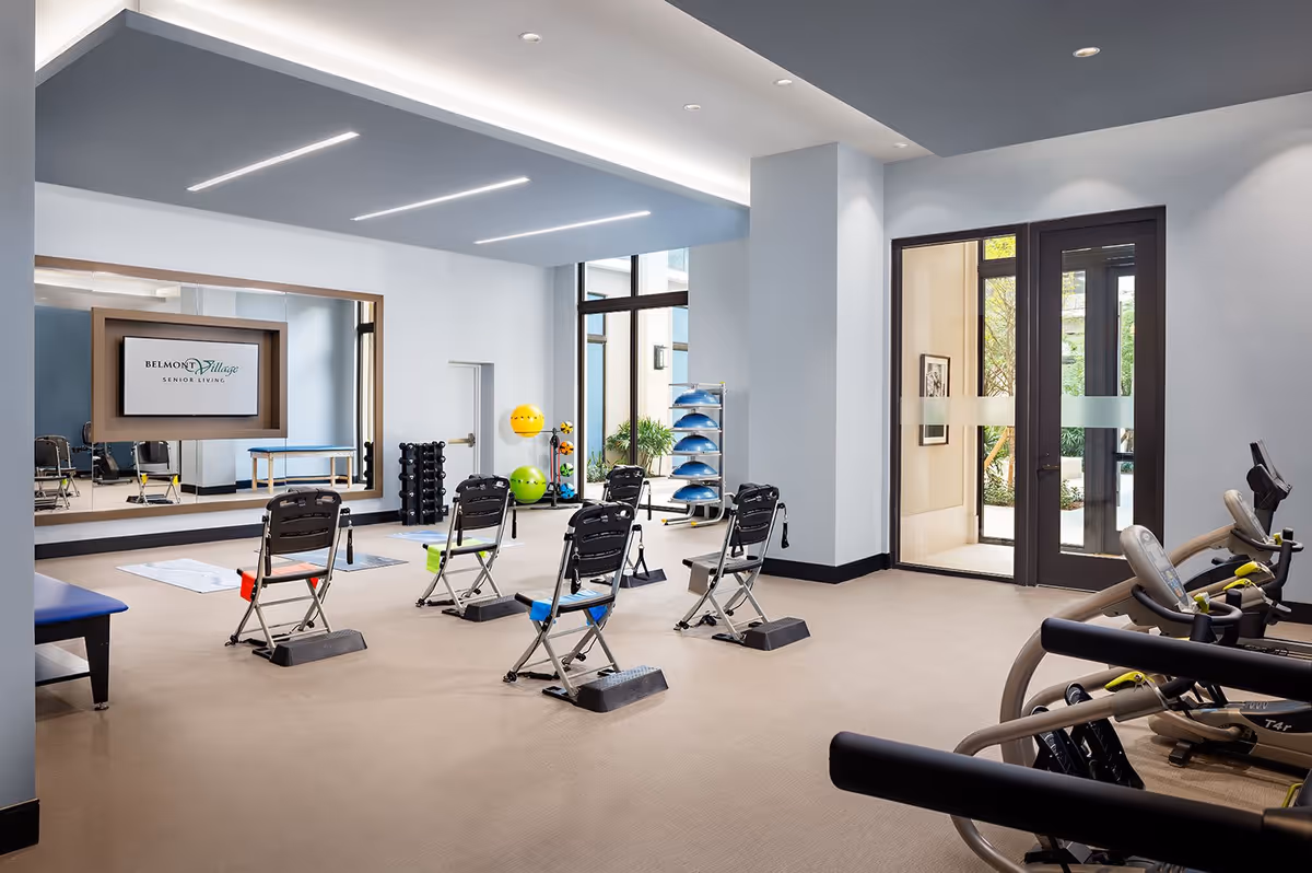 Bright fitness room with folding exercise chairs, cardio machines, stability balls, a mirrored wall, and a display reading Belmont Village Senior Living.