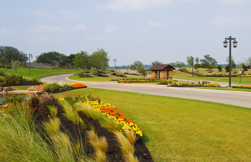 A landscaped outdoor area with a curved road, green grass, colorful flower beds, small trees, and lamp posts under a partly cloudy sky.
