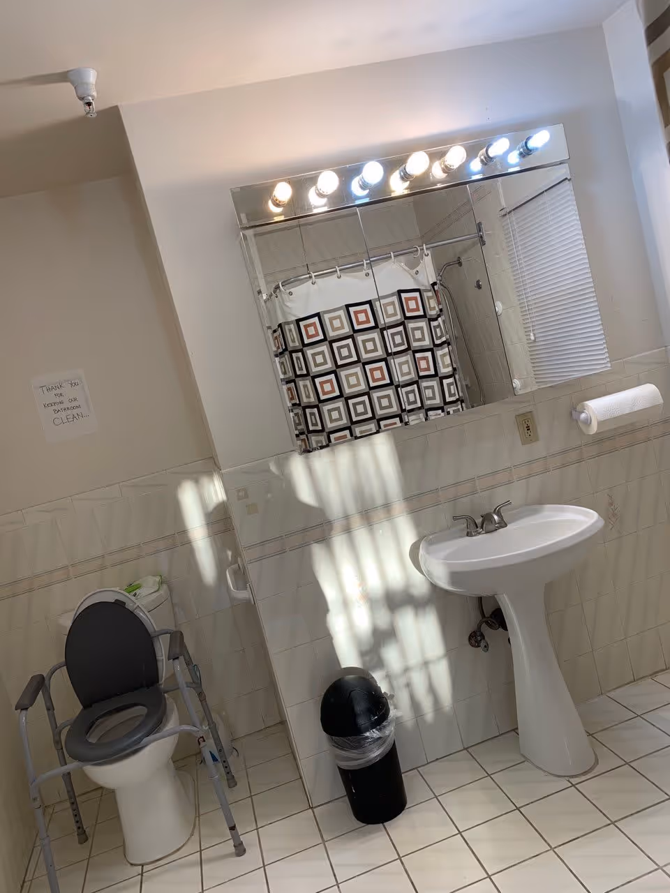 A clean bathroom with a white pedestal sink, a large mirror with multiple light bulbs above it, a toilet equipped with a gray safety frame, a black trash can with a plastic liner, a paper towel holder mounted on the wall, and a shower curtain with a geometric square pattern. There is a small sign on the wall that reads 'Thank you for keeping our bathroom clean.'