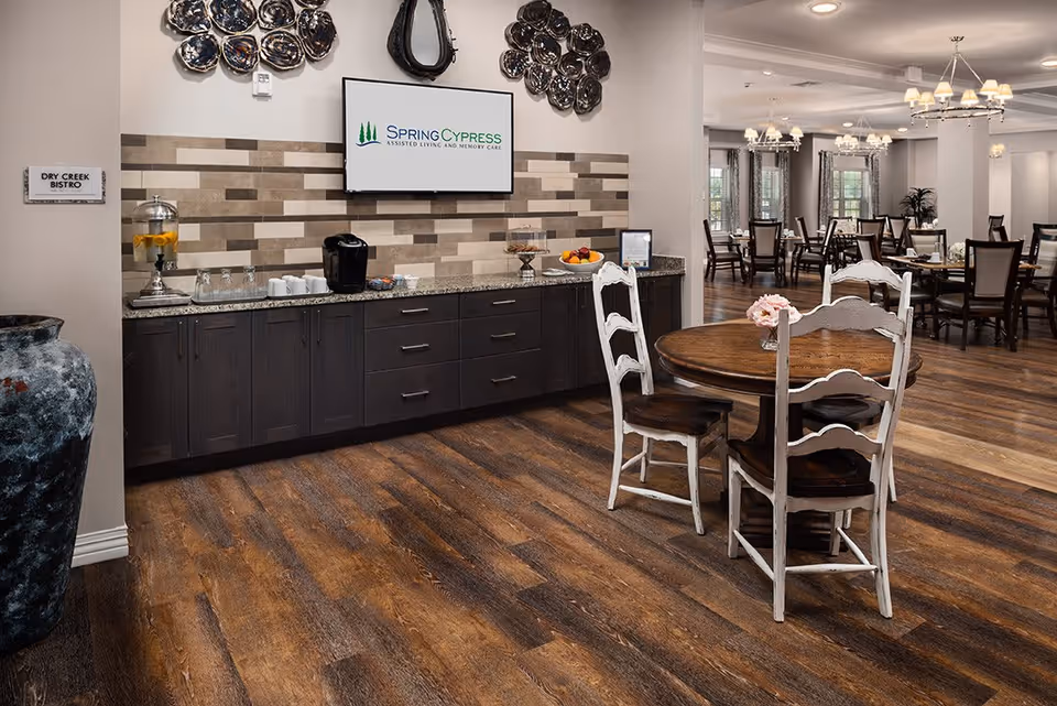 Interior view of a senior living facility dining area with wooden floors, a round wooden table with four white chairs, and a long counter with a coffee machine, glasses, and a fruit bowl. A wall-mounted screen displays the Spring Cypress Assisted Living and Memory Care logo. The room is well-lit with chandeliers and has multiple dining tables and chairs in the background.