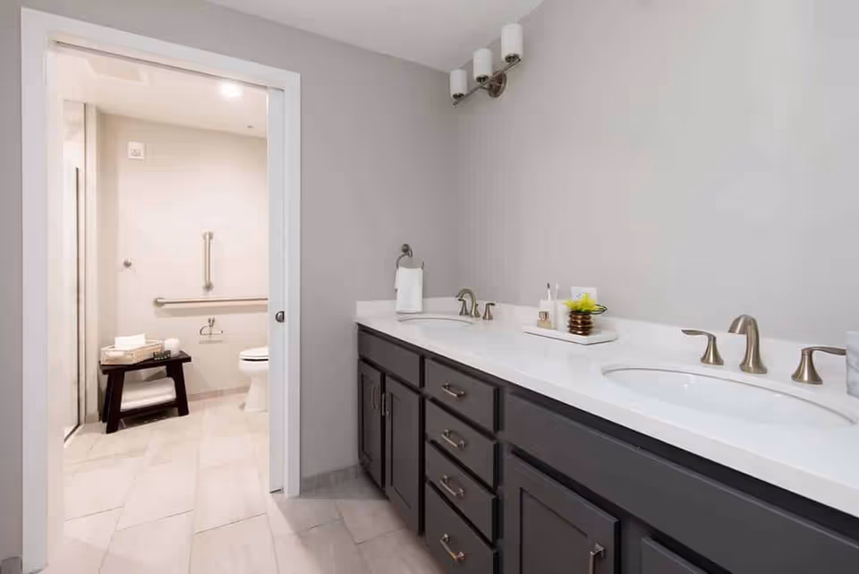 Modern bathroom with a long double-sink vanity, gray cabinets, and a doorway to a toilet and shower area.