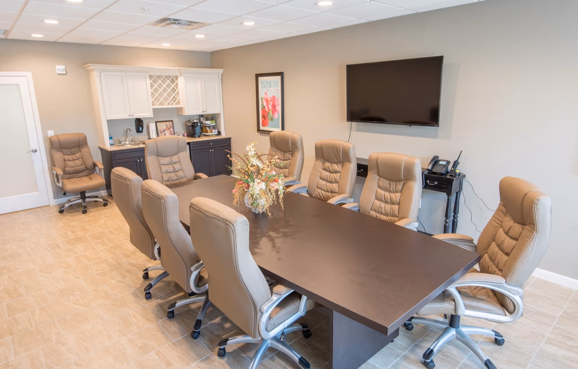 A conference room with a long dark wooden table surrounded by eight beige leather office chairs on wheels. A floral arrangement is placed in the center of the table. On the wall, there is a flat-screen TV mounted above a small black table holding a phone and a walkie-talkie. In the background, there is a kitchenette area with white upper cabinets, dark lower cabinets, a sink, a coffee maker, and a framed floral picture on the wall.