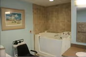 Interior view of a bathroom area in a nursing facility featuring a walk-in bathtub with tiled walls, a black chair with armrests, a scale, a framed picture on the wall, and a mirror reflecting part of the room.
