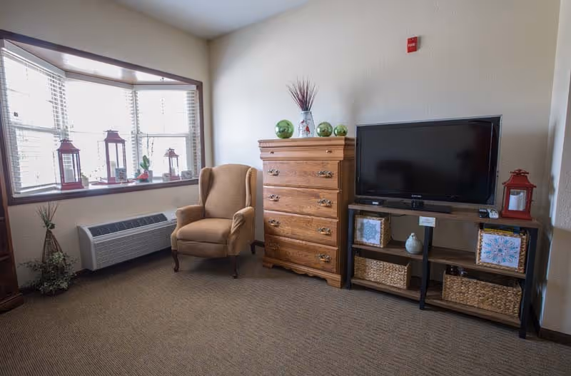 A cozy living room area with a beige armchair near a large window with white blinds. Next to the armchair is a wooden chest of drawers decorated with green glass spheres and a vase with dried flowers. A flat-screen TV sits on a black TV stand with woven baskets and decorative items on the shelves. The room has beige carpet and light-colored walls.