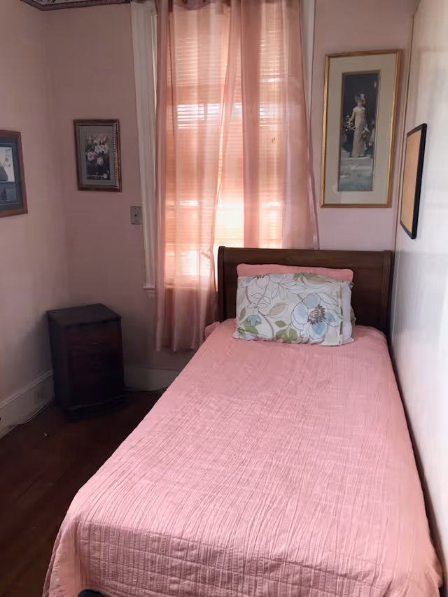 A small bedroom with a single bed covered in a pink quilt and a floral pillow. The bed is positioned against a wall with framed artwork hanging above it. A window with sheer pink curtains allows light to filter into the room. There is a small dark wooden nightstand next to the bed.