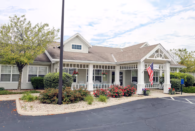 Exterior view of Taylor Place senior living facility showing a single-story building with a covered entrance, white columns, hanging flower baskets, an American flag, and landscaped bushes and flowers along the front. There is a parking area in front with a handicapped parking sign visible.