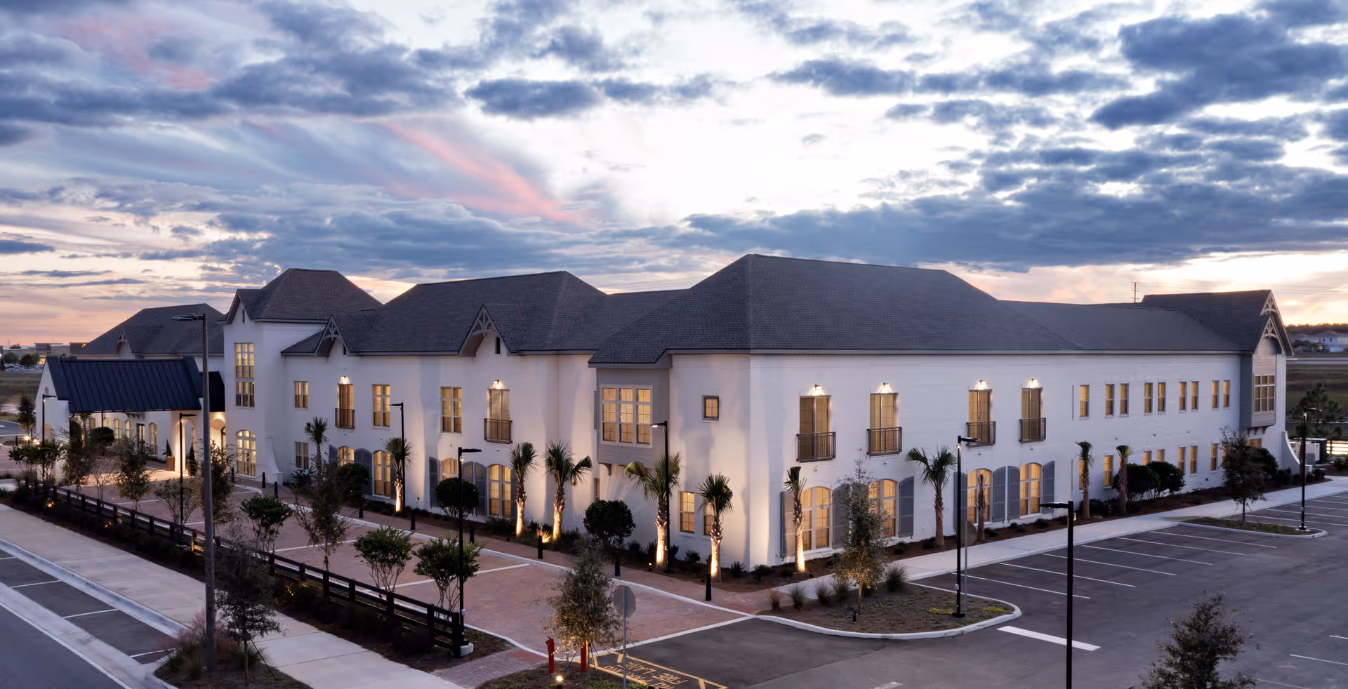 Exterior view of The Blake at Viera senior living facility at dusk, showing a large two-story building with multiple windows, palm trees, and a parking lot in front under a partly cloudy sky.