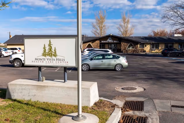 Exterior view of Monte Vista Hills Health Care Center showing the facility building, parking lot with several cars, a large sign with the facility name and logo, and a clear blue sky with some trees in the background.