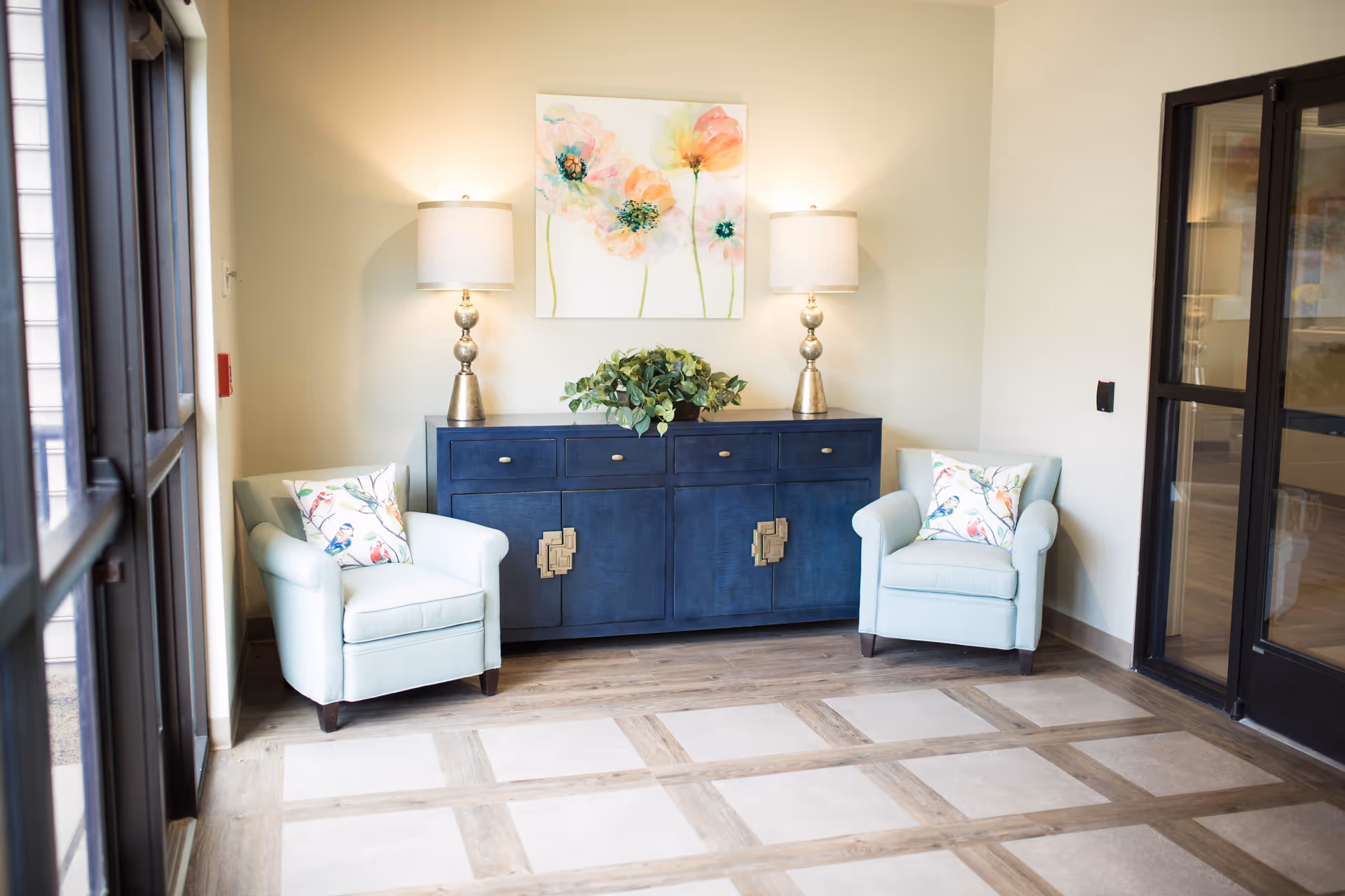 A cozy interior corner with two light blue armchairs, each with a decorative bird-patterned pillow. Between the chairs is a dark blue cabinet with four drawers and two doors, topped with two matching lamps and a green leafy plant. Above the cabinet hangs a colorful floral painting. The floor features a checkered pattern of wood and tile, and there are glass doors on either side of the space.
