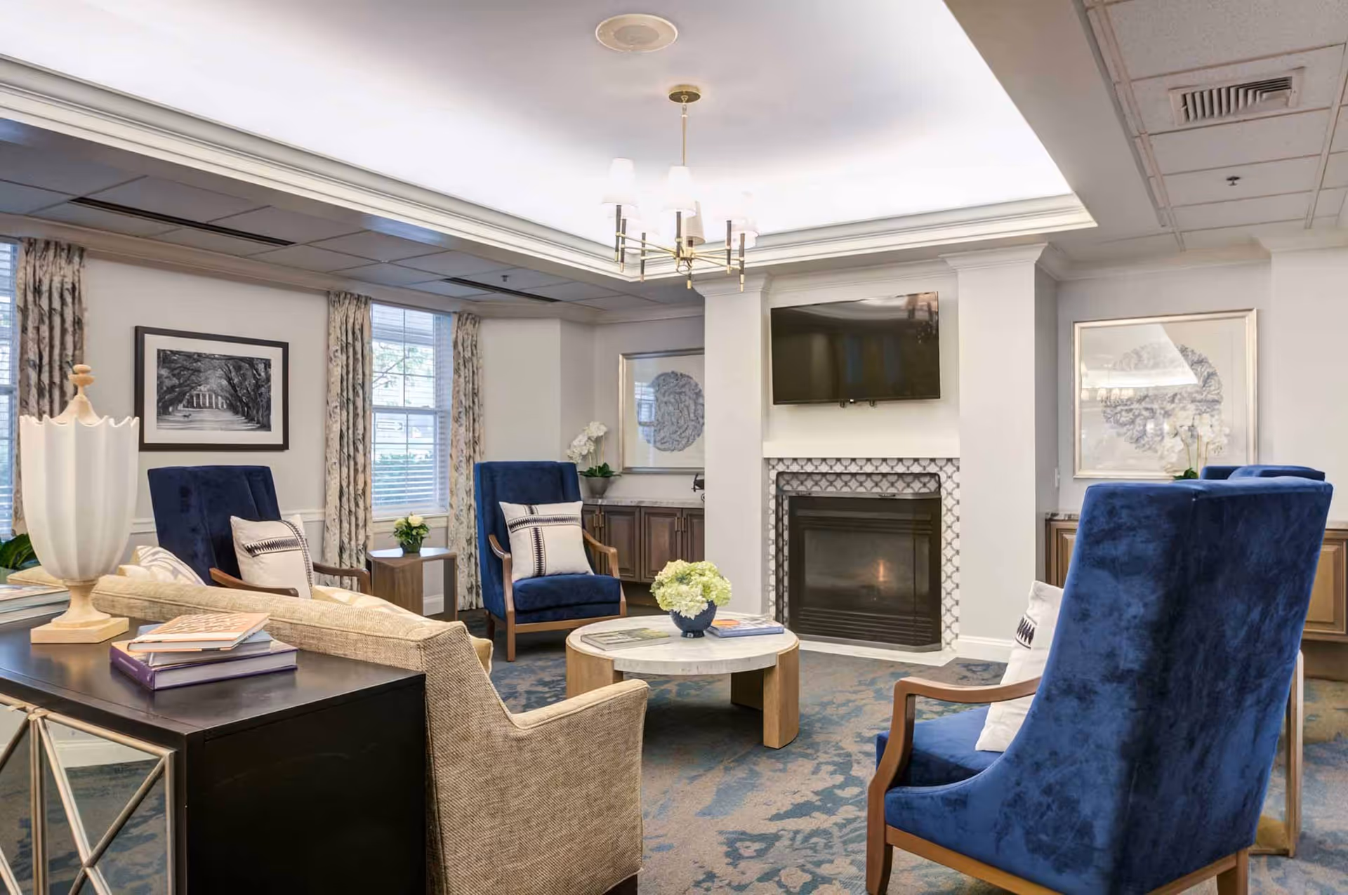 A cozy living room area with a beige sofa and three blue upholstered armchairs arranged around a round coffee table with a flower vase. The room features a fireplace with a mounted flat-screen TV above it, two framed artworks on the walls, windows with patterned curtains, and a ceiling light fixture.
