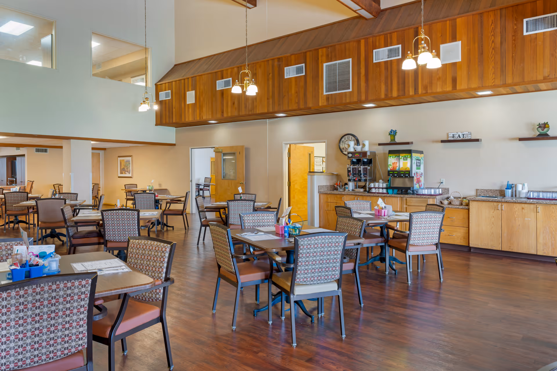 A spacious dining room with multiple tables and chairs arranged neatly. The room features wooden flooring, a high ceiling with wooden paneling, and hanging light fixtures. There is a beverage station with coffee and juice dispensers along one wall, and shelves with decorative items. The room appears bright and welcoming with an open doorway leading to another area.