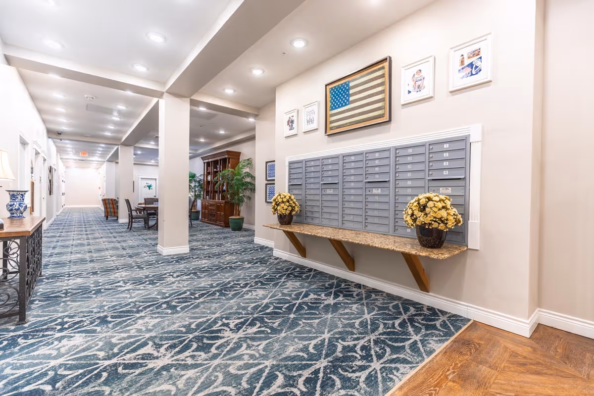 Bright carpeted hallway in an assisted living facility with a wall of mailboxes, decorative flowers, framed artwork, and seating areas.