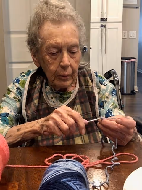 An elderly woman sitting at a wooden table, focused on knitting with blue and pink yarn. She is wearing a floral shirt and a plaid apron, with a wheelchair visible behind her. The background shows a white door, cabinets, and a trash can in a well-lit room.