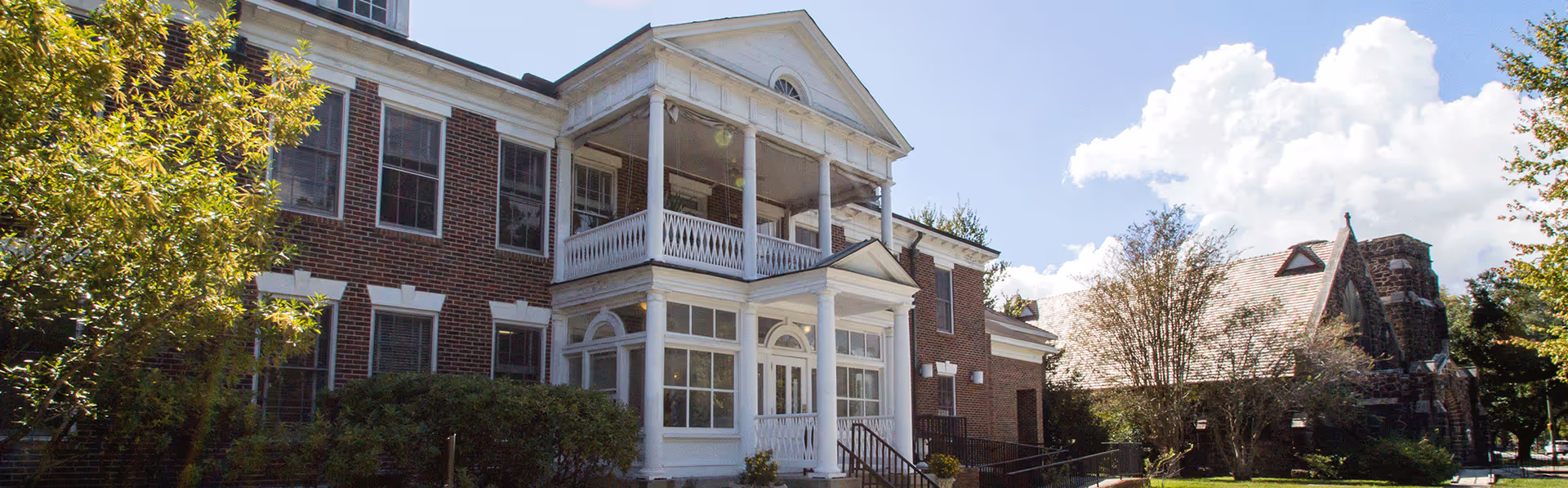 Exterior view of a two-story brick building with white columns and a balcony, surrounded by trees and bushes under a partly cloudy sky.