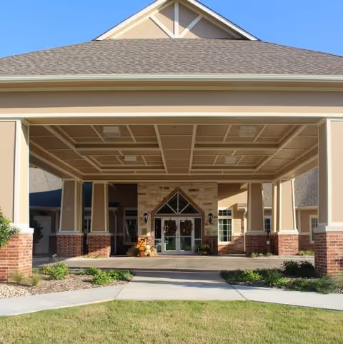 Entrance of a senior living facility with a covered driveway supported by beige columns with brick bases, a triangular window above double glass doors, and some autumn decorations near the entrance.