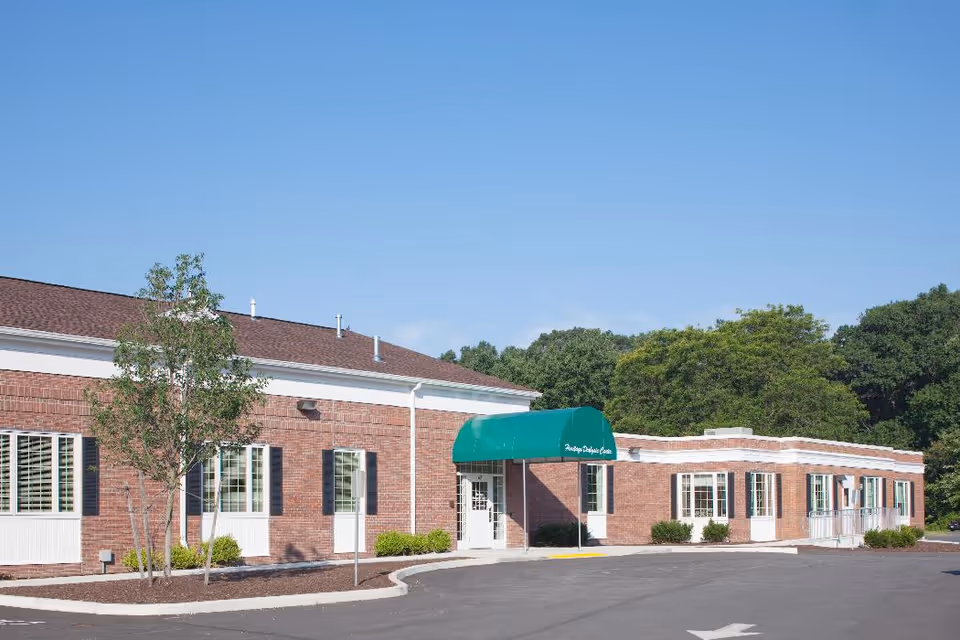 Exterior view of a single-story brick building with white trim and a green awning over the entrance. There are several windows with black shutters, a small tree, and shrubs planted in front of the building. The sky is clear and blue.