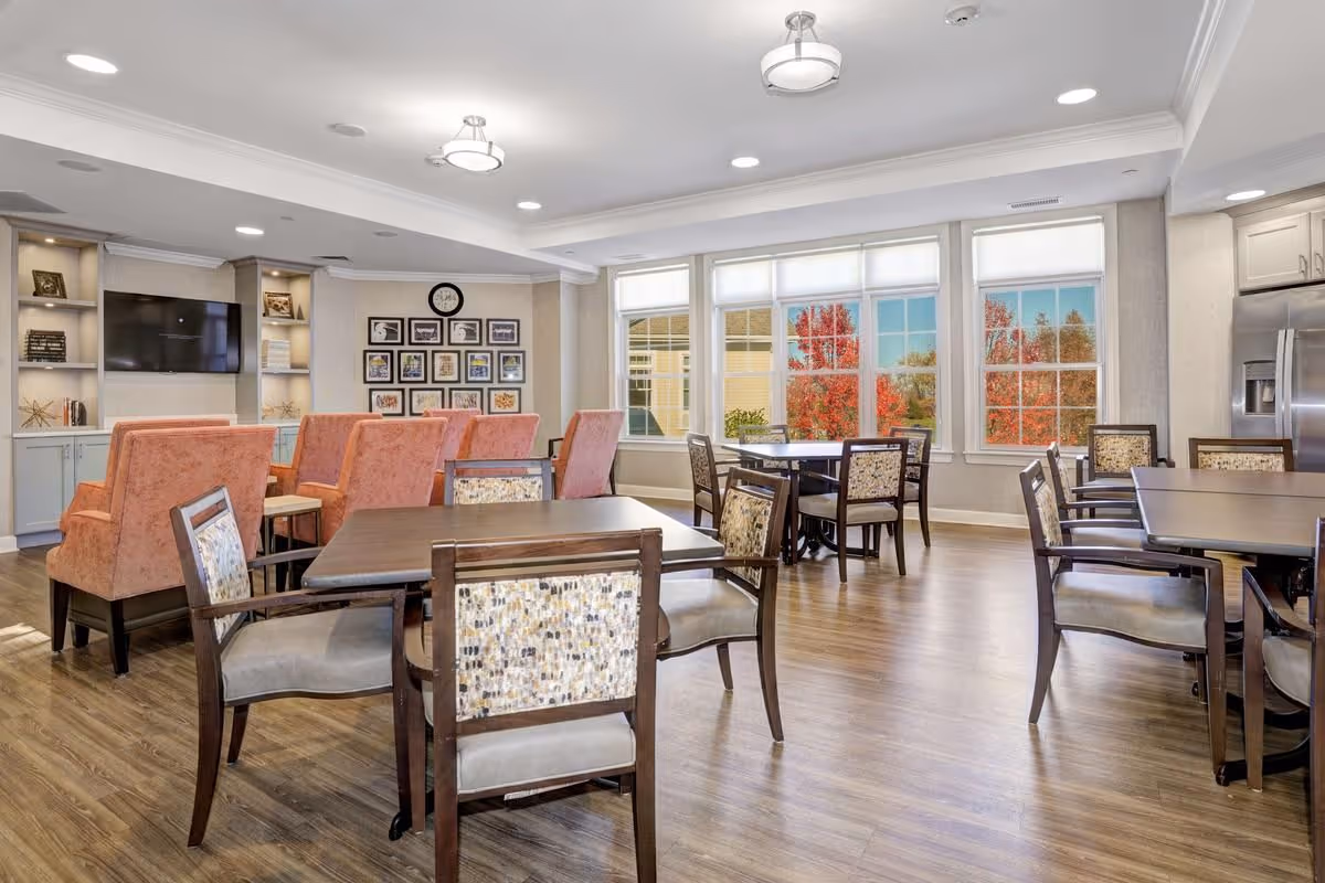 Bright communal dining and lounge area with tables, chairs, upholstered armchairs, a TV, and large windows showing autumn foliage.