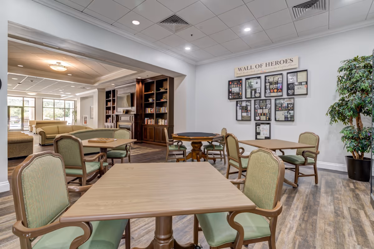 A spacious common area in a senior living facility featuring several tables with green cushioned chairs. In the background, there is a cozy seating area with beige sofas, a wooden bookshelf, and a fireplace. On the right wall, a 'Wall of Heroes' display showcases framed memorabilia. The room has wood flooring and a ceiling with recessed lighting.