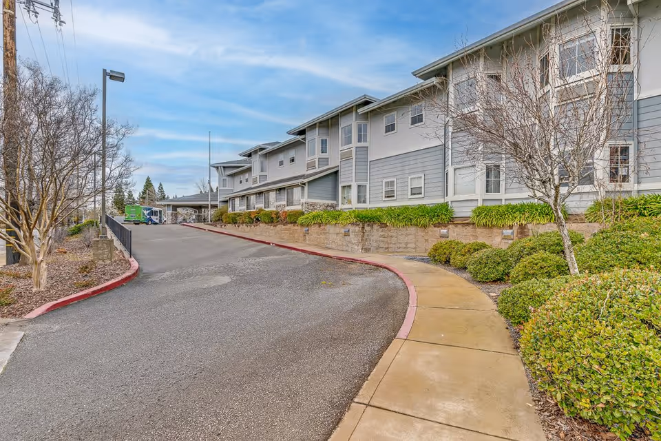 Exterior view of a multi-story senior living facility building with gray and white siding, surrounded by landscaped bushes and trees. A curved sidewalk runs alongside the building, and a driveway leads up to the entrance. The sky is partly cloudy.